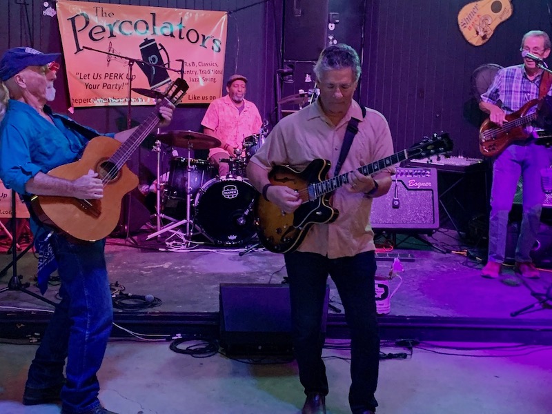 A four-member band, The Percolators, is performing on stage. The guitarist on the left is wearing a hat and blue shirt, another guitarist in front is in a beige shirt, the drummer at the back sports a pink shirt, and the bassist on the right wears a checkered shirt. A banner reading "The Percolators" is visible in the background.