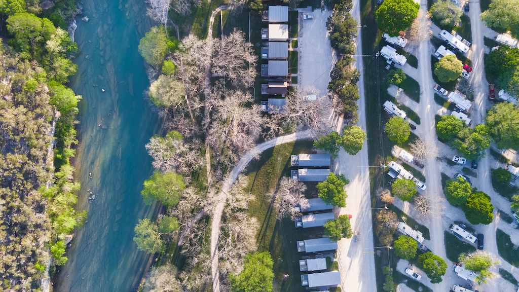 Aerial view of Rio Guadalupe flanked by campsites and cabins nestled among trees. The clear water flows on the left side, while the right side shows multiple neat rows of RVs in an RV park and a curved road. Patches of grass and lush foliage create a serene, nature-filled setting.
