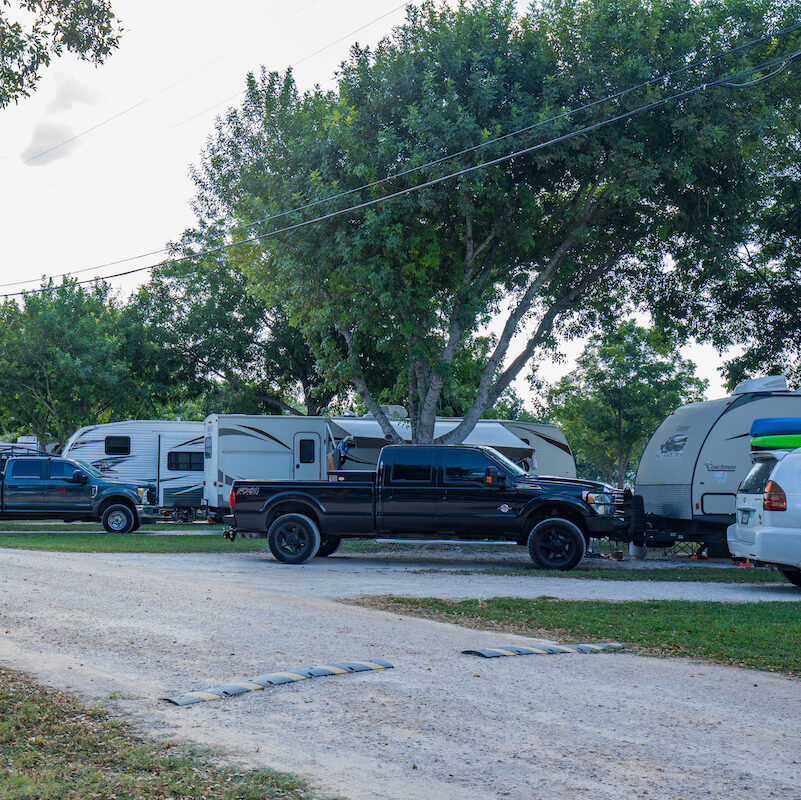 A tranquil scene at Rio Guadalupe RV Park shows several parked RVs and trailers under large trees. A black pickup truck stands in front, towing a trailer. Other vehicles and trailers dot the grassy areas along a gravel road beneath a clear sky.