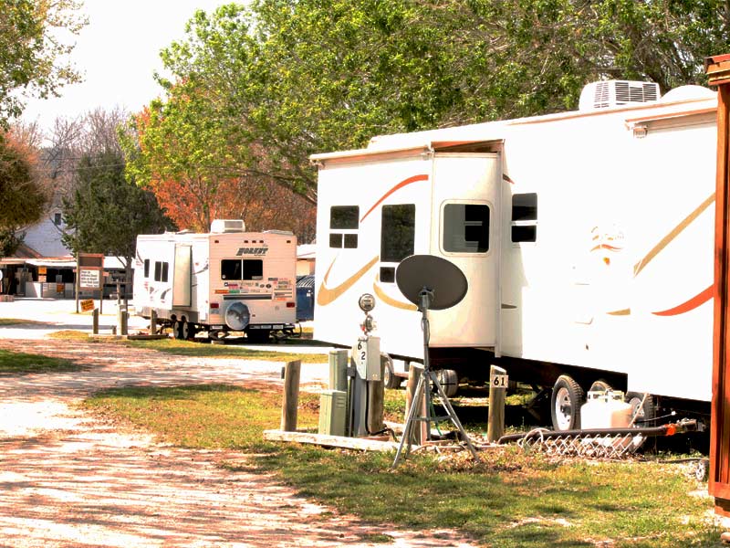 Several recreational vehicles (RVs) are parked in the landscaped Rio Guadalupe RV Park with hookups. The scene includes trees and a gravel pathway, with one RV showing a satellite dish and other utilities connected. The setting is calm, suggesting a serene outdoor environment.