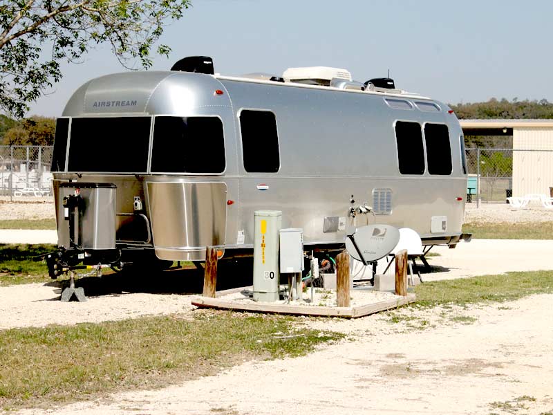 A shiny silver Airstream trailer is parked on a grassy campsite at Rio Guadalupe RV Park. It is attached to utilities, including an electrical box and a satellite dish. The background shows a wire fence, some trees, and a light-colored building under a clear sky.