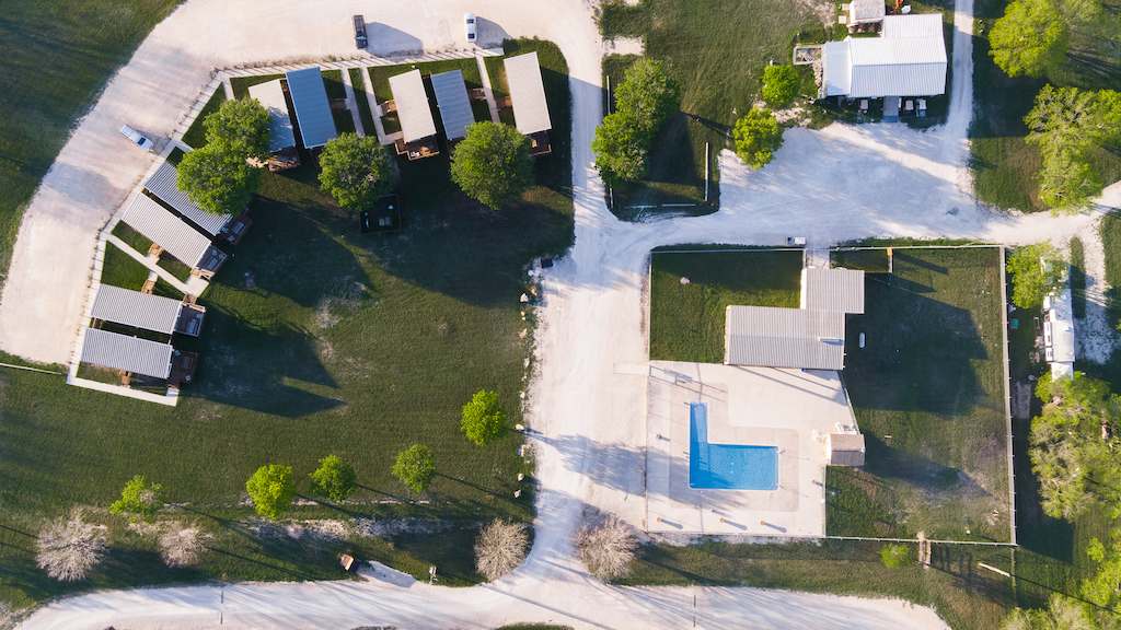 Aerial view of Rio Guadalupe RV Park campground with cabins arranged in a curved line on the left. A rectangular swimming pool is visible in the center-right, surrounded by grass and trees. Various paths and roads connect the cabins and pool area, forming a clear layout.