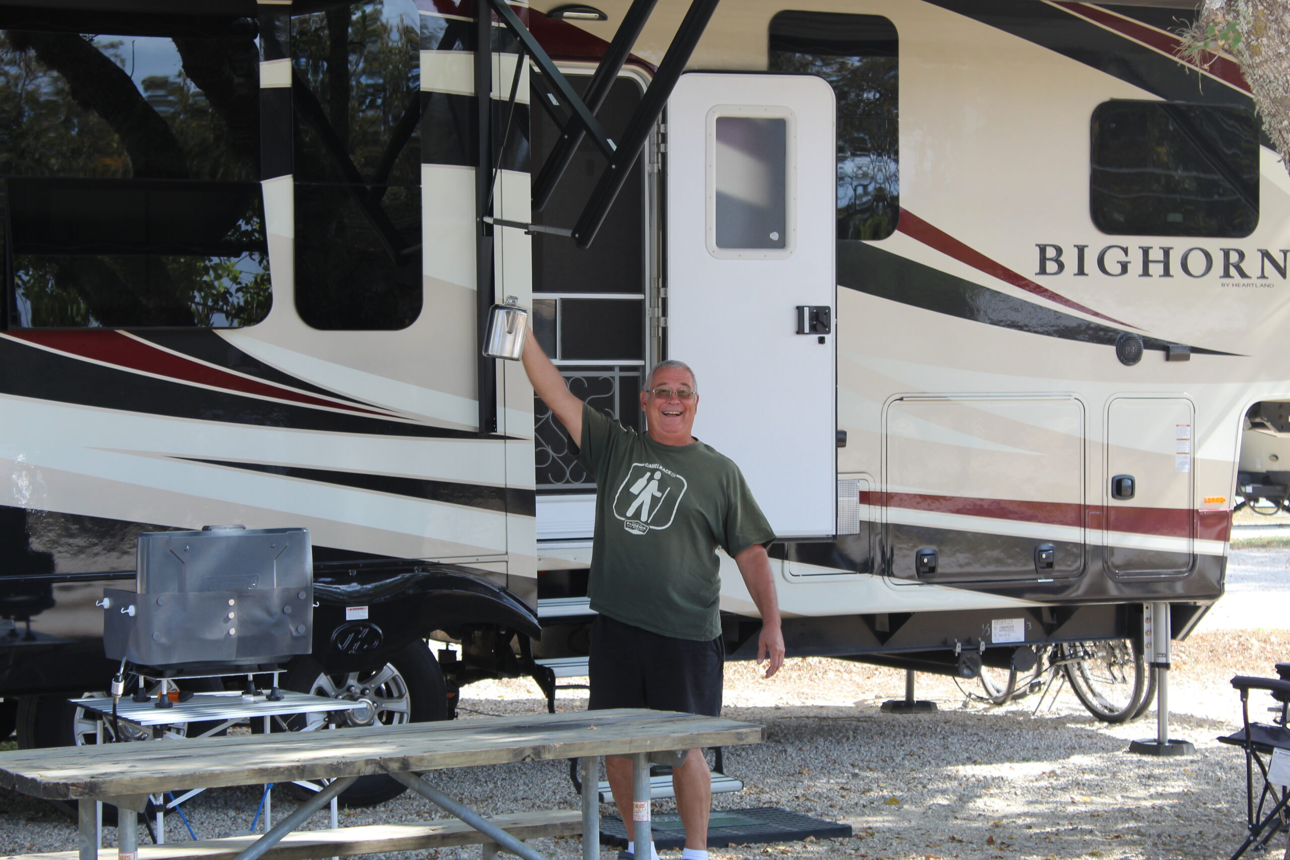 A man stands outside a Bighorn RV, smiling and holding onto the entry handrail. He's wearing a green t-shirt, glasses, and has grey hair. In the foreground, there's a wooden picnic table and a portable grill on a metal stand, adding to the cozy home feel of his outdoor setup.