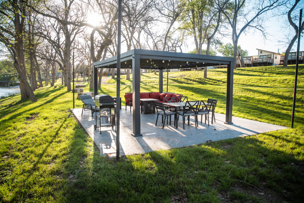 A covered outdoor seating area with a rectangular pergola, grilling station, and dining table set on a concrete slab, surrounded by green grass and trees. Sunlight filters through the trees, and a quiet river runs nearby with buildings visible in the background.