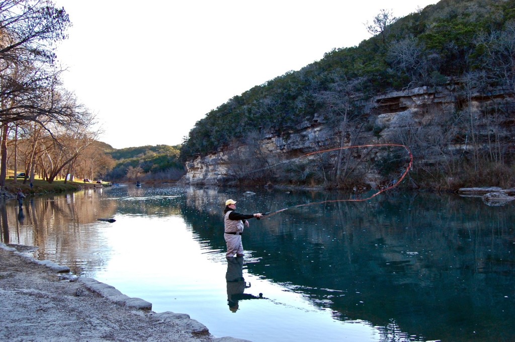 A person stands knee-deep in the calm, reflective waters of the Guadalupe River, casting a line with a large circular motion. The river, flanked by rocky cliffs and tree-lined banks, is perfect for fly fishing under the clear sky.