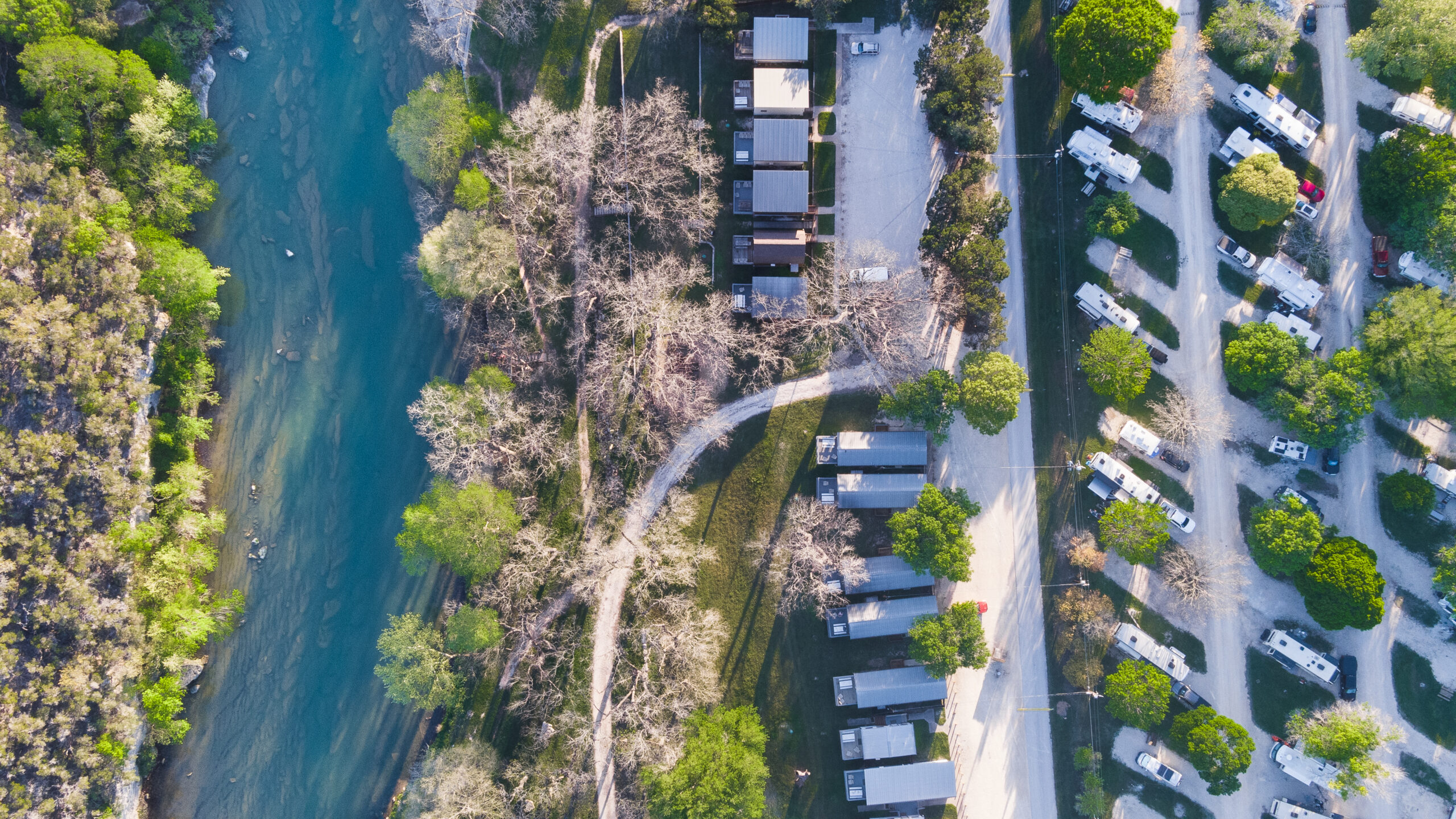 Aerial view of a riverside campground with several small, rectangular cabins arranged in a semi-circular layout near a winding river. Adjacent to the cabins, RVs and trailers are parked in rows among trees, offering shade and greenery to the camping area—perfect for your main page.