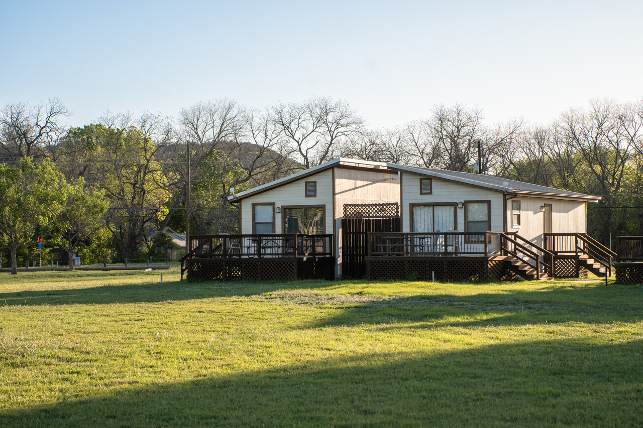 A single-story home with a large wooden deck sits in a grassy open field. Surrounding the property are numerous bare and leafy trees under a clear blue sky, basking in sunlight. The house features multiple stairs leading up to the deck.