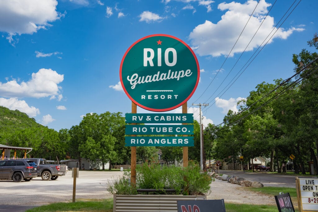 A large green and red circular sign for Rio Guadalupe Resort advertises RV & cabins, Rio Tube Co., and Rio Anglers, with trees, parked cars, and a blue sky with clouds in the background.