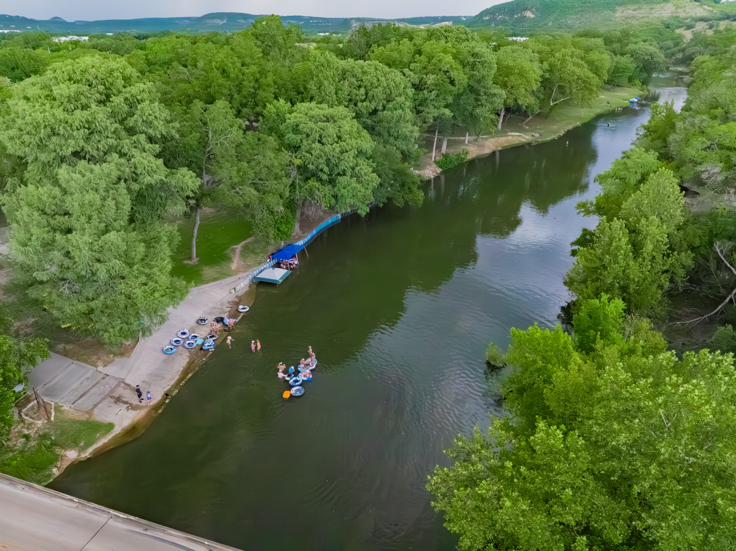 A group of people relax on inner tubes and swim in a calm river surrounded by lush green trees, with a ramp and some gear visible on the riverbank. The landscape is hilly in the distance.