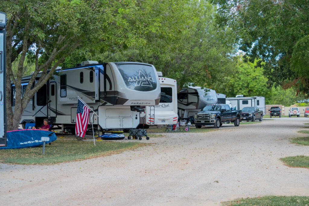 Rows of RVs and trucks are parked along a gravel road at a campsite surrounded by green trees. An American flag stands near one RV, and outdoor chairs and a table are set up beside it.