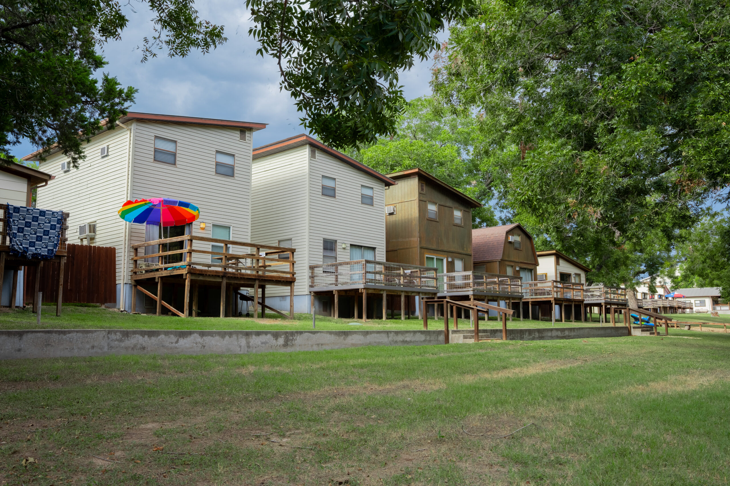 Row of elevated lakeside cabins with wooden decks, one featuring a colorful umbrella. Large green trees provide shade, and a grassy lawn stretches in front of the buildings under a partly cloudy sky.