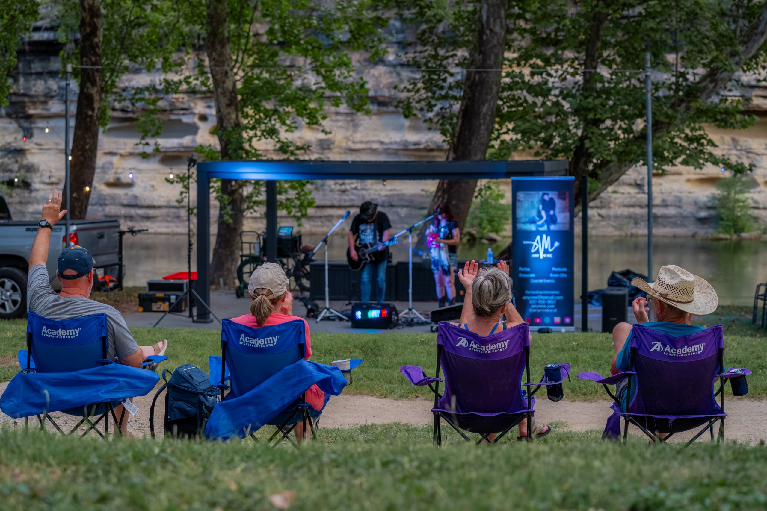 Four people in folding chairs watch an outdoor band perform on a small stage near trees and a rocky riverbank. Some audience members clap and raise their hands, enjoying the live music on a summer day.