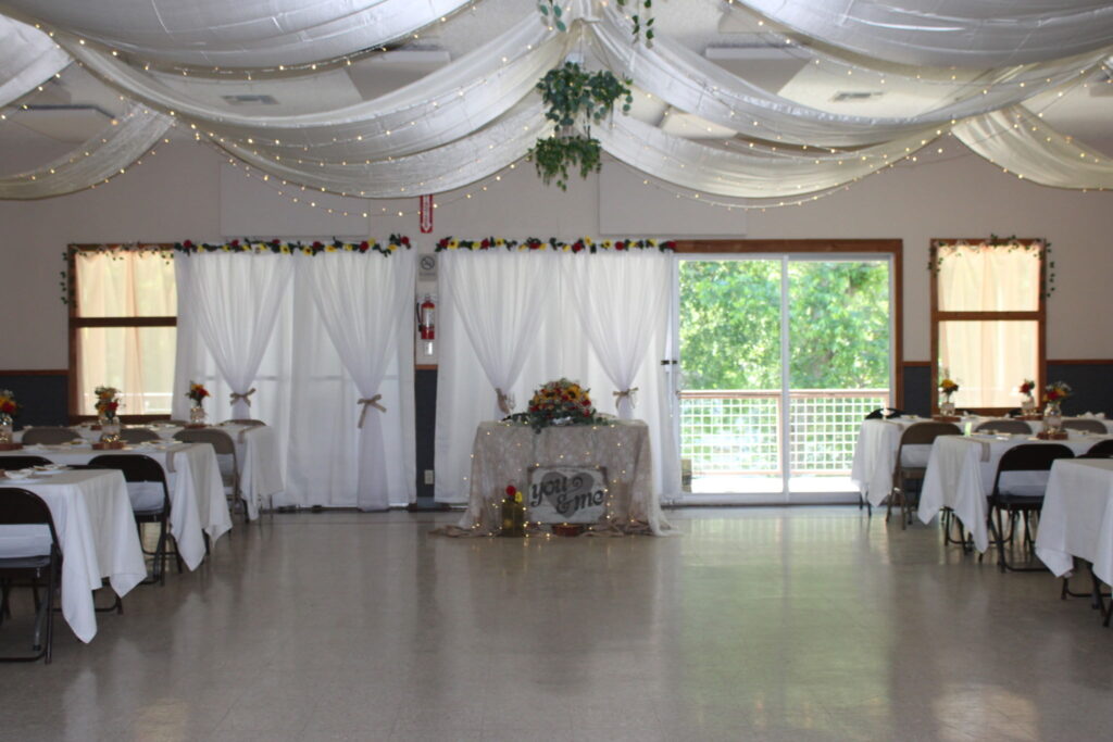 A decorated event hall with tables covered in white cloths, floral centerpieces, and a main table with a "Love" sign. White drapes and string lights hang from the ceiling, with sunlight streaming through windows and sliding doors.