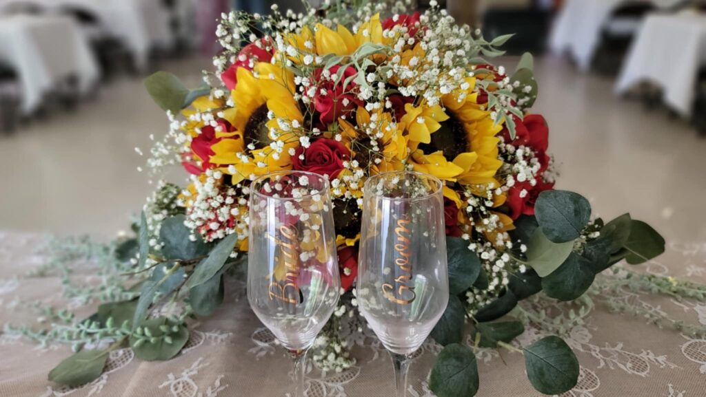 Two champagne glasses labeled “Bride” and “Groom” stand in front of a bright floral arrangement featuring sunflowers, red roses, greenery, and baby's breath on a lace-covered table.