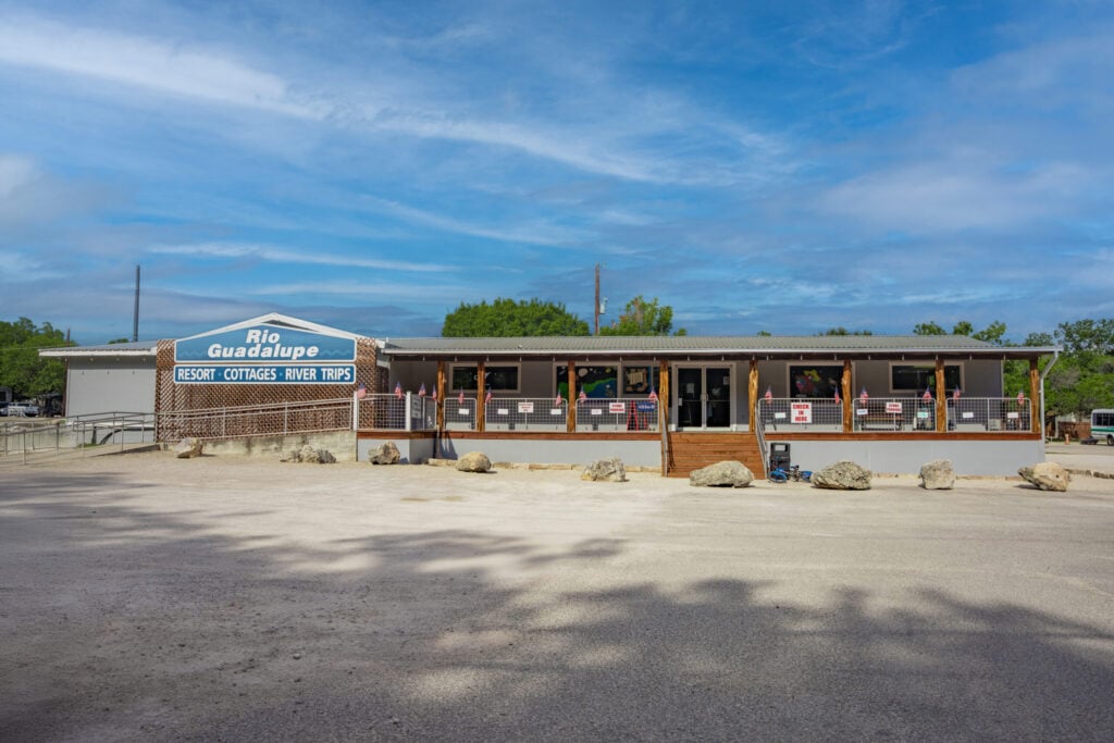 A single-story building with a sign reading "Rio Guadalupe Resort Cottages River Trips" stands under a blue sky, with a wide porch, several chairs, and rocks scattered across the gravel parking lot in front.