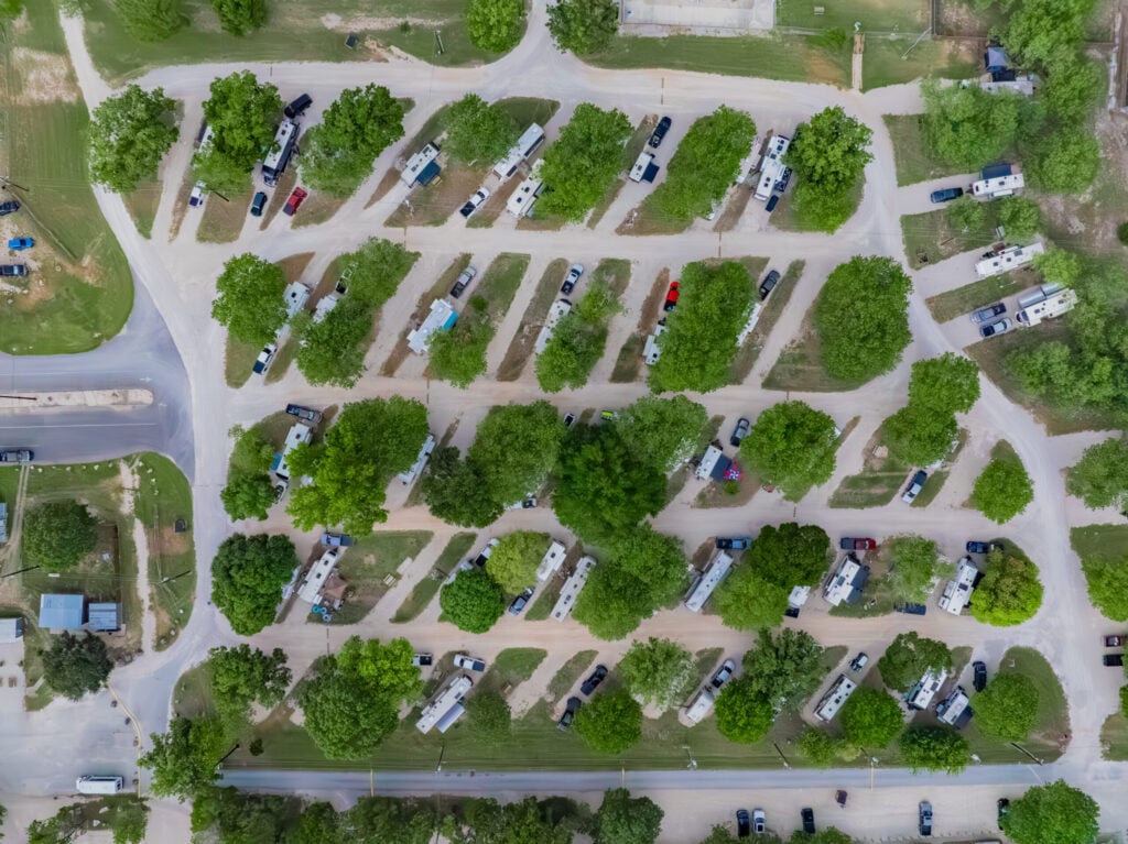 Aerial view of an RV park with rows of parked RVs separated by green trees and grassy areas. Several cars are parked next to the RVs, and paved roads run through the park. The surrounding area is also green and lush.
