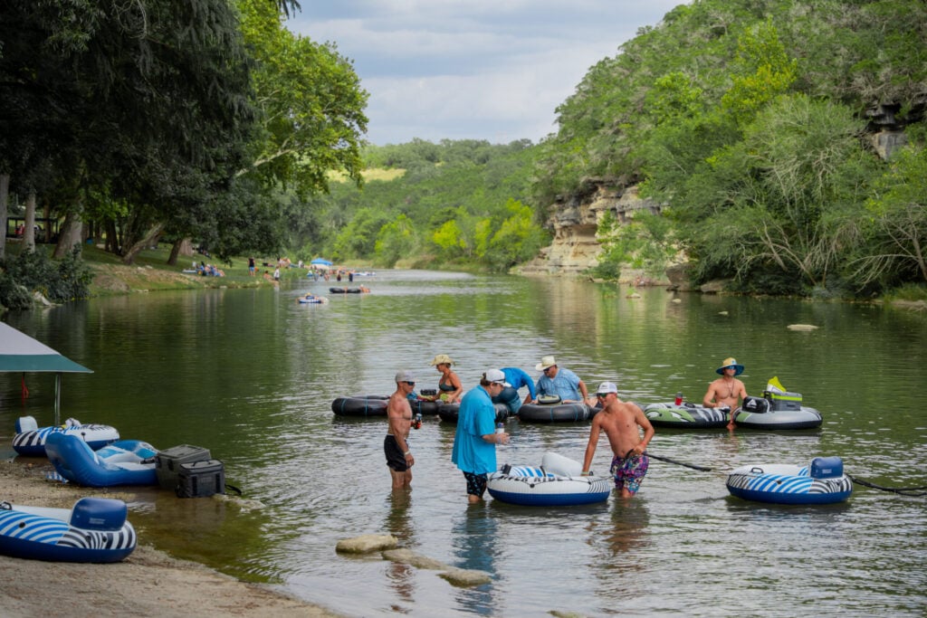 People relax on inflatable tubes in a calm river surrounded by greenery and rocky cliffs. Some stand in the shallow water while others float nearby under a partly cloudy sky.