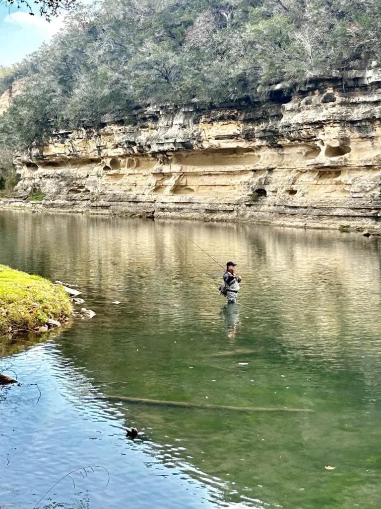 A person stands waist-deep in a calm river, fly fishing near a rocky cliff with layered, weathered stone and trees on top. The water reflects the cliff and surrounding greenery under a bright sky.