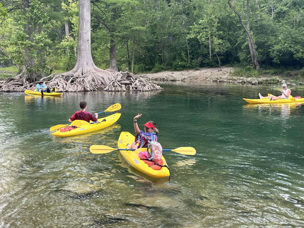 People in bright yellow kayaks paddle down a calm, green river surrounded by trees. One person waves while others relax or paddle, and a tree with exposed roots is visible on the riverbank.