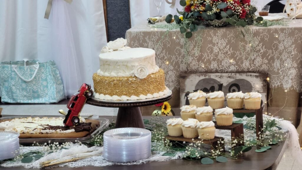 A decorated dessert table with a white and gold two-tier cake, a small toy fire truck, several white-frosted cupcakes, plastic plates, and floral arrangements on a lace tablecloth.