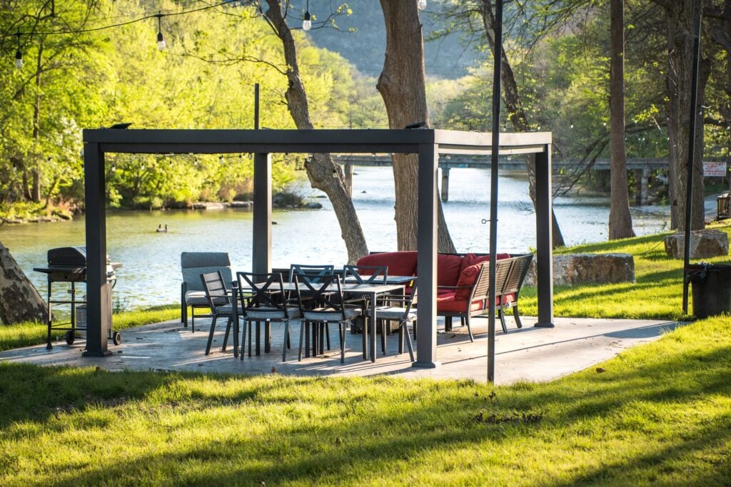 A modern outdoor patio with a pergola, dining table, chairs, and red sofa set beside a river. Surrounded by green trees and grass, the area is bright and inviting, with string lights and a grill nearby.