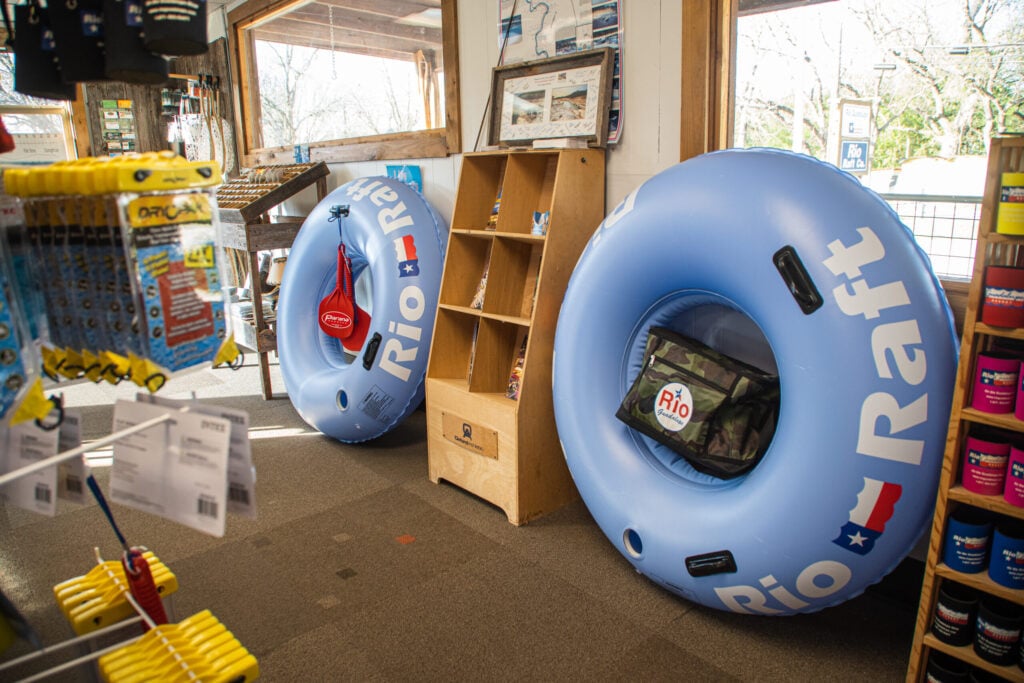 Two large blue Rio Raft inflatable tubes with handles and pockets are displayed upright inside a shop, surrounded by shelves with various merchandise. Sunlight streams in through large windows.