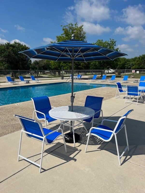 Outdoor swimming pool area with blue lounge chairs and a white round table under a large blue umbrella—perfect for relaxing after water sports or enjoying sunny days surrounded by green trees and clear skies.