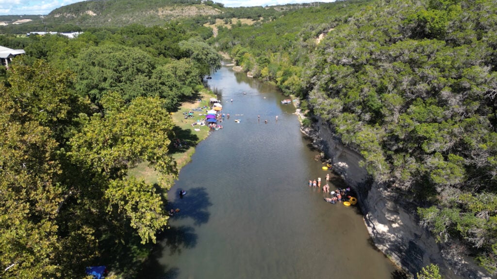 Aerial view of a river winding through a forested area with people enjoying river activities—swimming, relaxing on the banks, and floating on colorful inflatables under sun umbrellas.