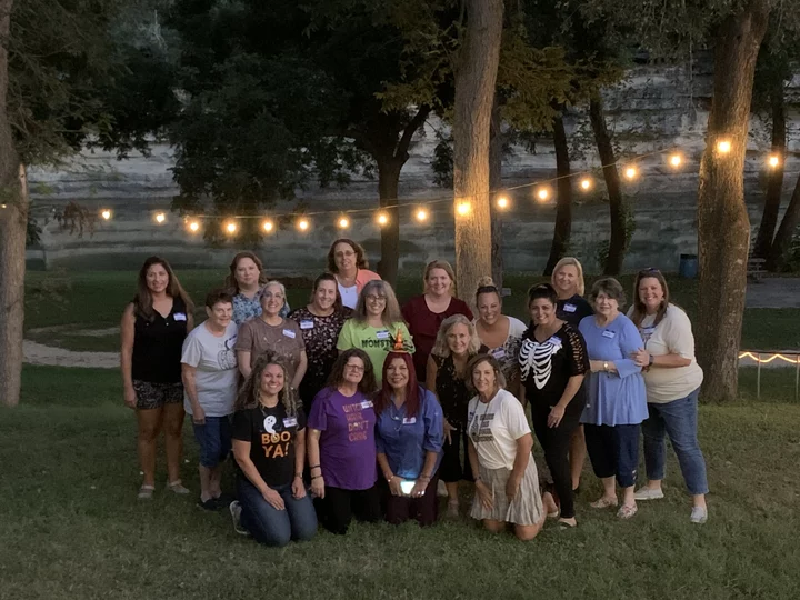A group of women poses together outdoors in the evening under string lights, surrounded by trees and grass, with some wearing casual clothes and a few in themed shirts.