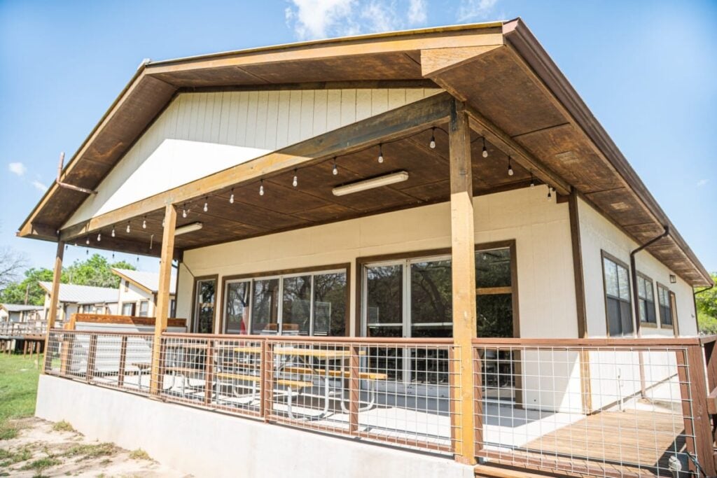 A modern cabin with a large covered porch, string lights, railings, picnic tables, and sliding glass doors, set in a grassy area with trees and blue sky in the background.