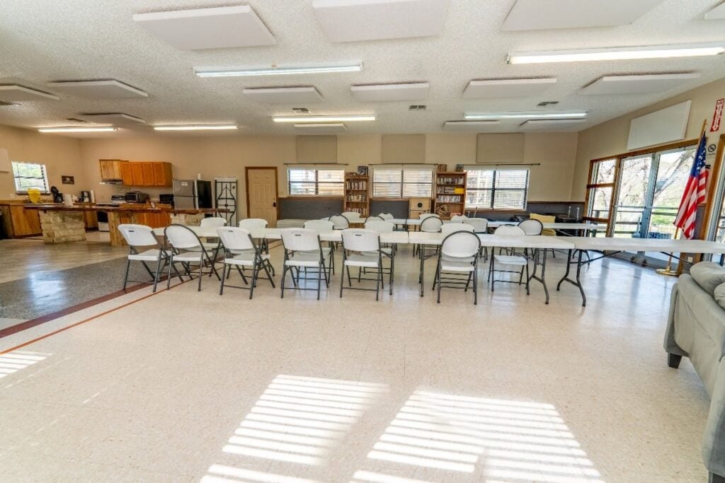 A bright, spacious community room with folding tables and chairs arranged in rows, a kitchen area on the left, bookshelves at the back, and large windows letting in natural light. An American flag stands on the right.