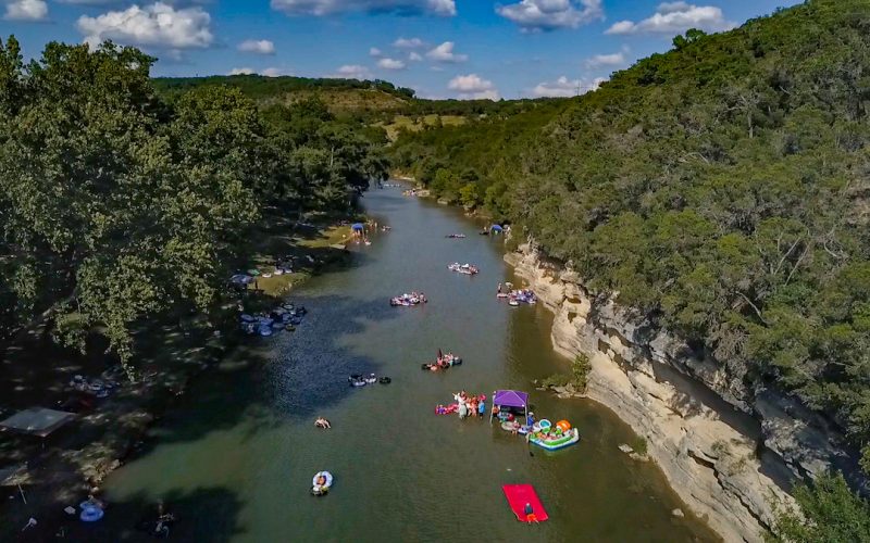 Aerial view of the scenic Guadalupe River surrounded by lush greenery and hills. The river is dotted with people enjoying tubing and rafting on inflatable tubes and rafts. Some groups are gathered under canopies on the riverbank, while a colorful floating platform is visible in the water.