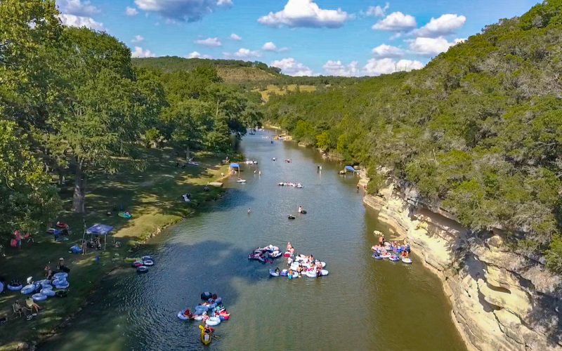 A scenic river surrounded by lush trees and rolling hills with scattered clouds overhead. People are enjoying recreational activities in and along the Guadalupe River, including rafting, tubing, and swimming. Some are relaxing on inflatable tubes and floats.