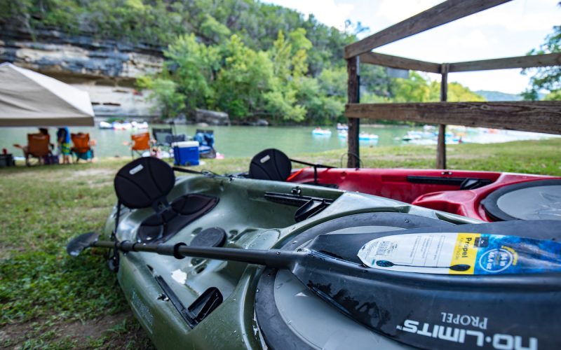 Several kayaks are gathered on a grassy area near the calm Guadalupe River, surrounded by lush green trees. A wooden railing and a tent are visible nearby, and in the distance, more kayaks and people can be seen enjoying kayak rentals on the sunny water.